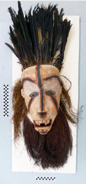 Clay human-like face mask with black and red striped accents. Feathers and course fibers make up the hair, beard, and headdress. 