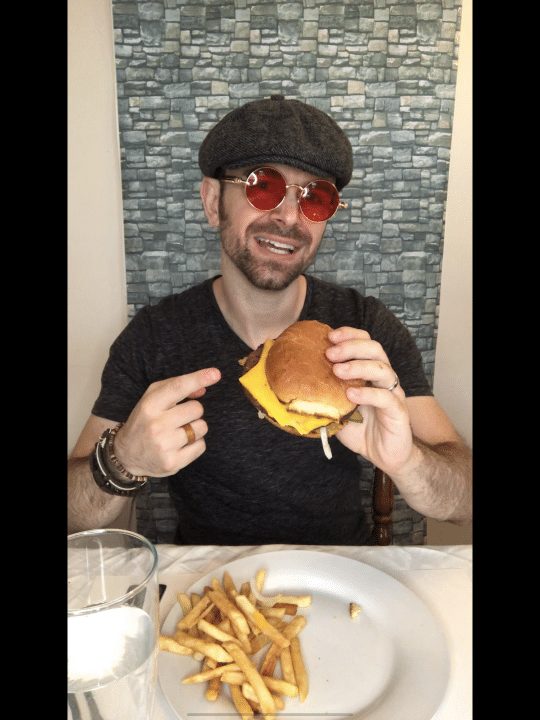 man wearing pink glasses and a hat holding a cheeseburger