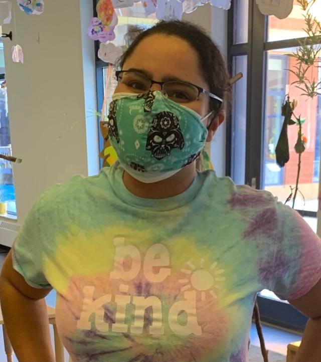 woman standing in front of a window wearing a face mask and a tie-dye t-shirt that says be kind