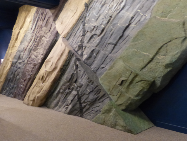 Benedum Hall of Geology strata wall. Shows different colors of rock stratigraphy from left to right: tan, blue-grey, maroon, beige, dark gray, olive green. 