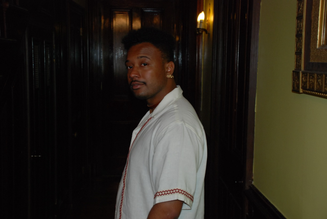 Black man in a white collared shirt standing in front of a wooden door indoors. 