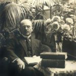 Black and white photo of a man in a suit holding a book. He is surrounded by books and plants.