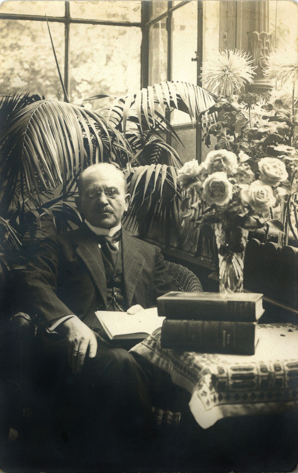 Black and white photo of a man in a suit holding a book surrounded by books and plants. 