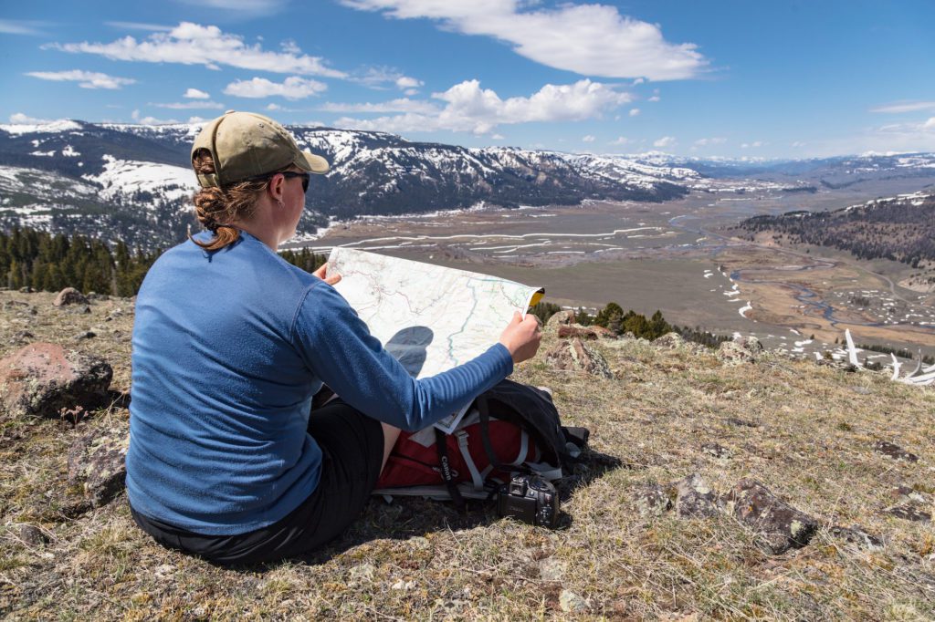A person looking at a park map is seated on a mountainside overlooking a river valley with snow capped mountains in the background