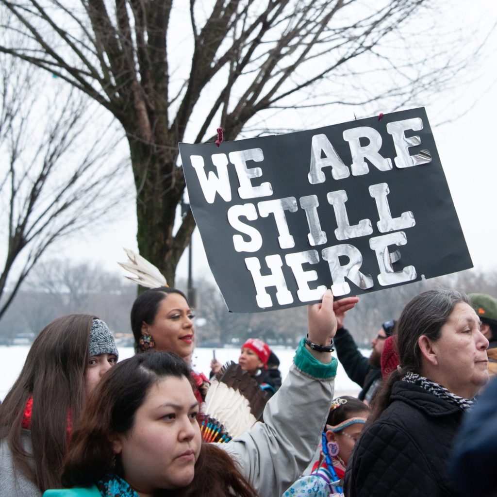 Indigenous person holding a sign that says "We Are Still Here"